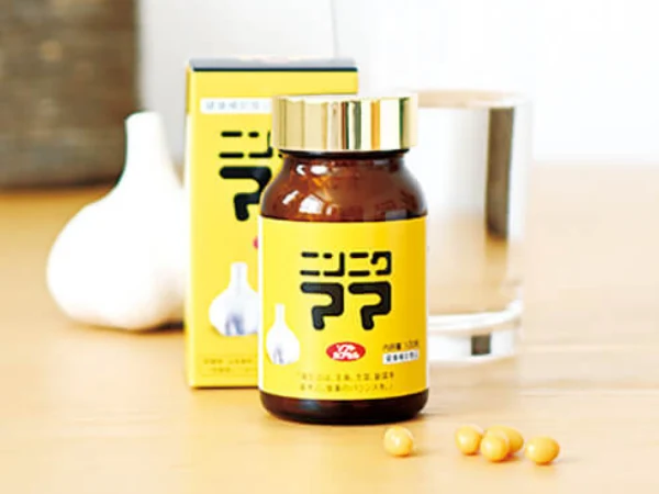 アイスター商事「ニンニクママ（サプリメント）」の商品イメージ、茶色の小瓶、濃い黄色のラベルに「ニンニクママ」と商品名の表記とにんにくのイラスト、金色のキャップ。背景にはニンニクと黄色い商品小箱、ソフトカプセル、透明なガラスのコップ。
